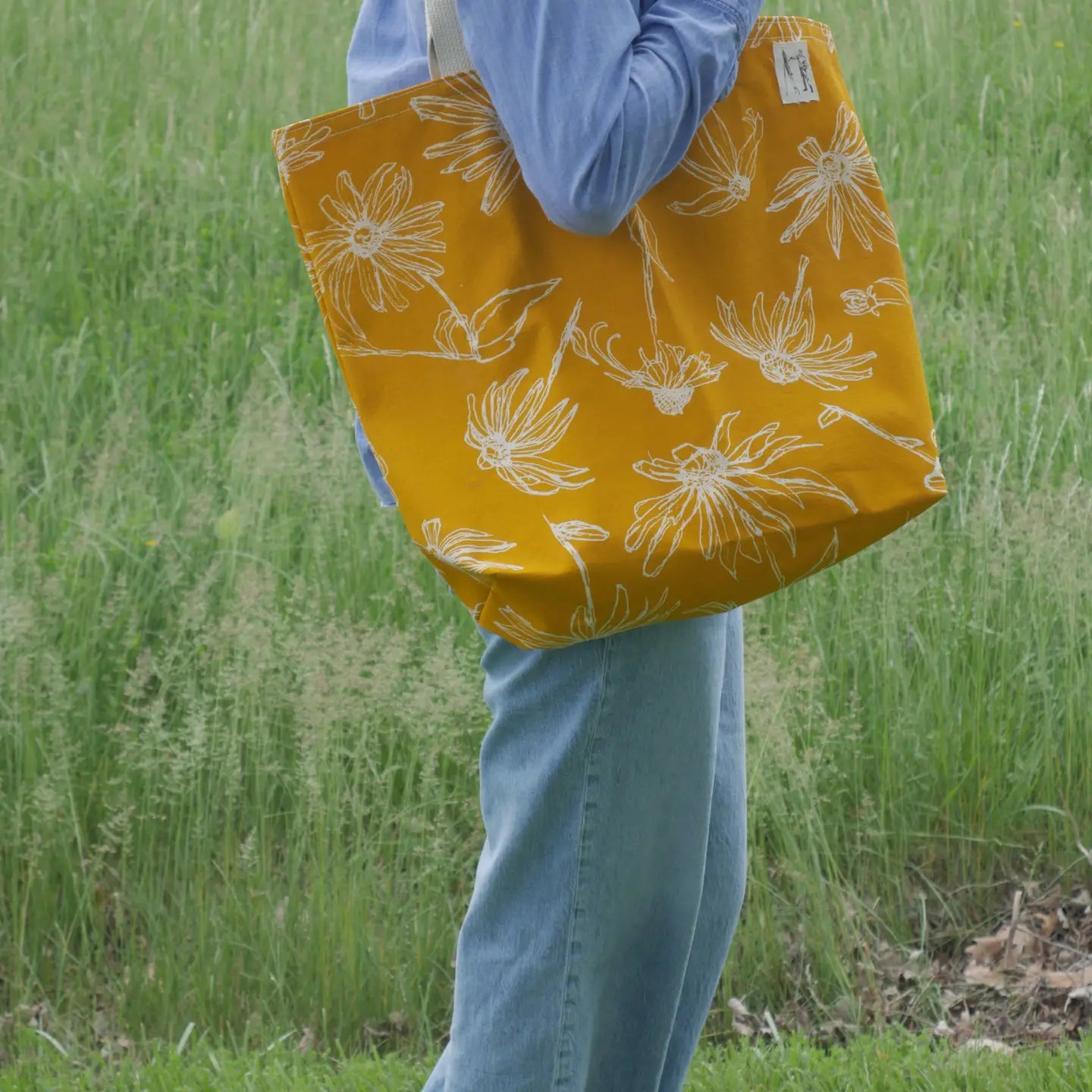 Person holding a yellow tote bag with floral pattern in a grassy field