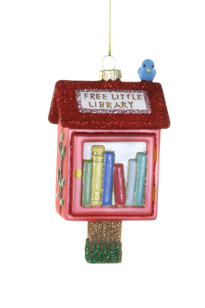 A glass ornament shaped like a free little library with red sides, a red glittered roof and a blue bird sitting on top. Suspended from a gold hanging cord shown on a white background.