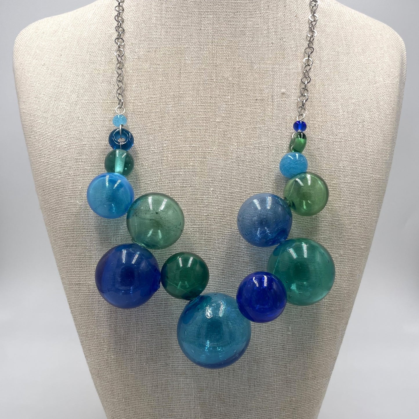 A chunky statement necklace with translucent glass beads in shades of blue, teal, and green, suspended from a silver chain and displayed on a beige fabric mannequin bust.