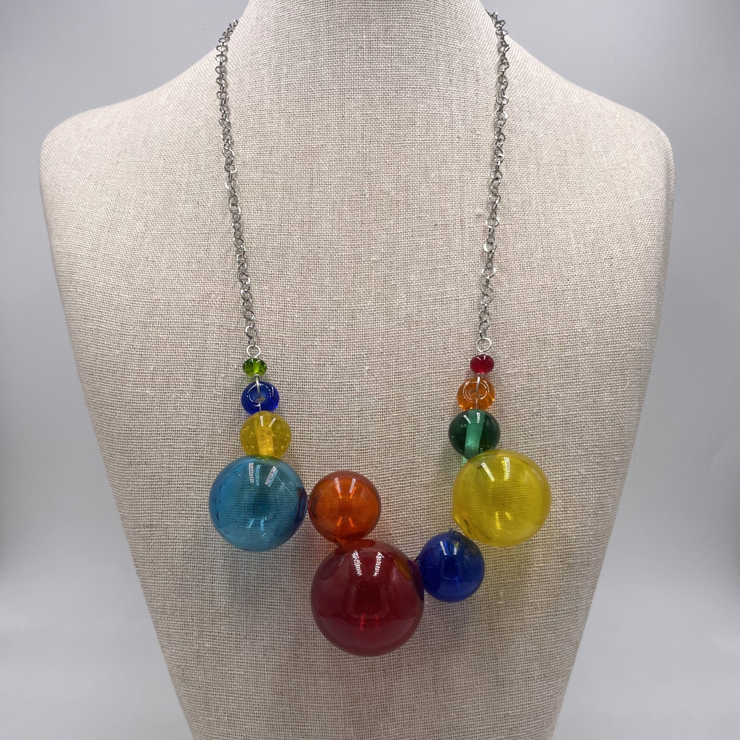 A colorful glass bead necklace displayed on a beige fabric mannequin bust. The necklace features a silver chain and a cluster of translucent glass beads in blue, red, yellow, orange, and green.