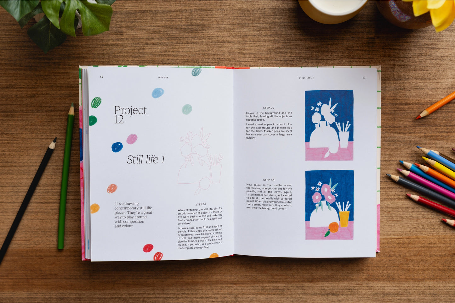 An open book laid flat on a wooden desk, showing printed pages with text and small colorful illustrations. Surrounding the book are colored pencils, with some green leaves and a cup visible at the top edge.