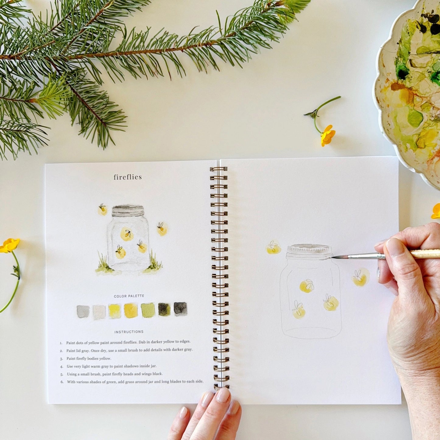 Person holding a paintbrush over an open book with art instructions, surrounded by nature-themed decor.