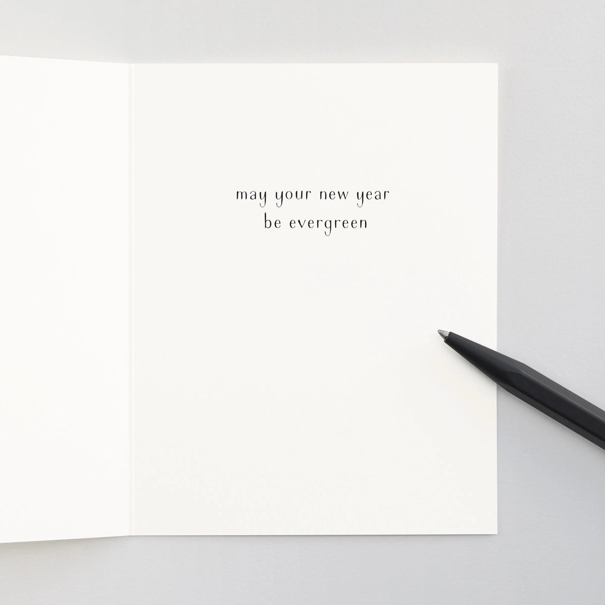 An open white notecard with the text "may your new year be evergreen" on a ligt grey background with a black pen.