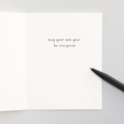 An open white notecard with the text "may your new year be evergreen" on a ligt grey background with a black pen.