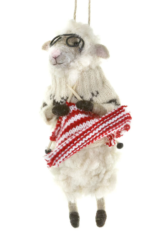 A decorative wool sheep shaped ornament wearing glasses and a sweater knitting a red and white stripe scarf. Suspended from a tan cord and shown on a white background.