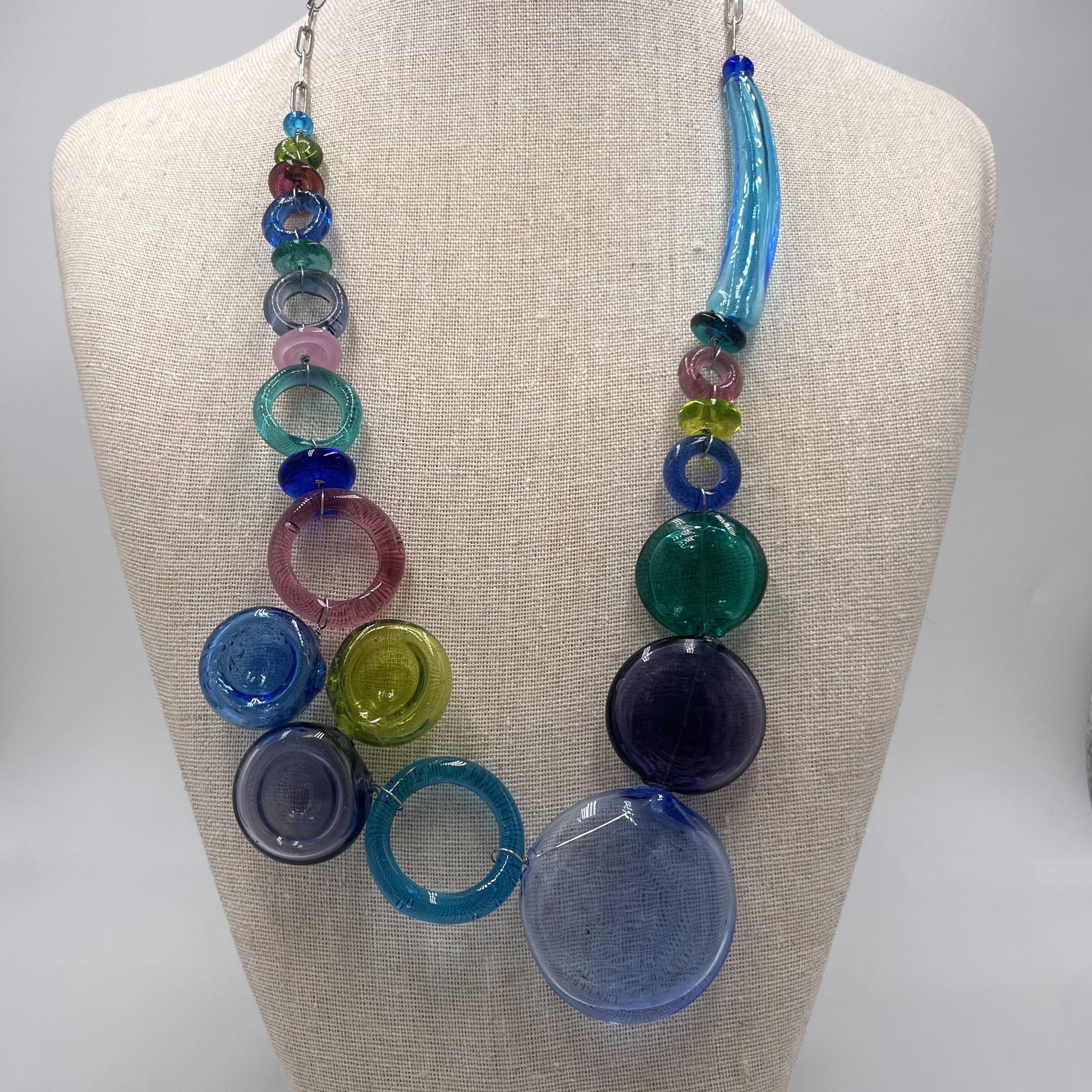 A colorful glass beaded necklace displayed on a beige fabric jewelry bust. The necklace features a mix of translucent round and cylindrical beads in shades of blue, green, pink, purple, and yellow.