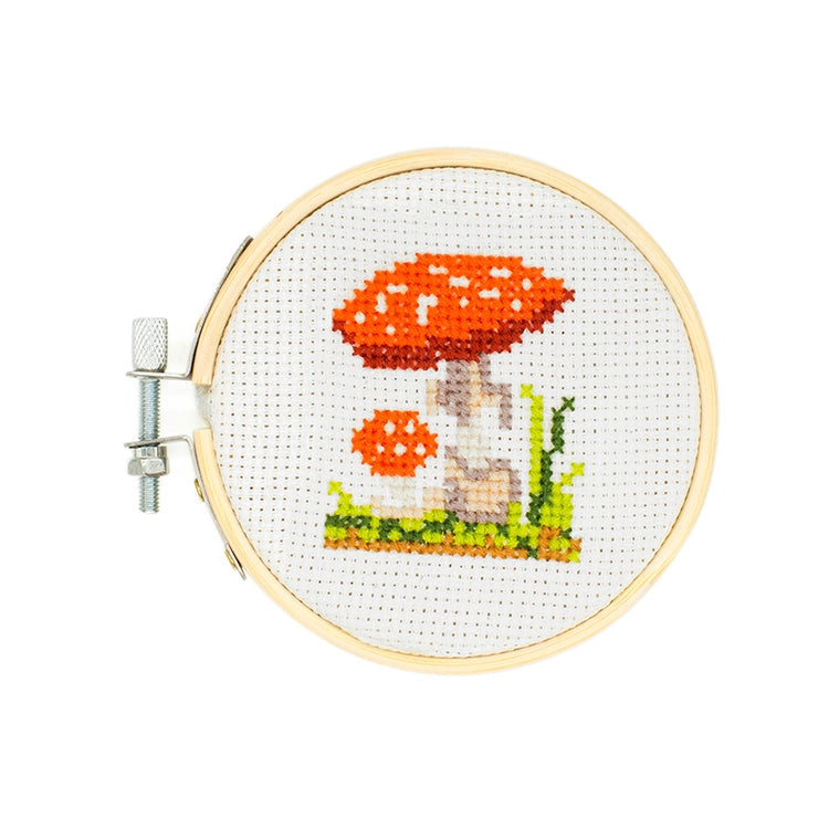 A completed cross-stitch embroidery in a wooden hoop, featuring a red mushroom with white spots,  green grass, and a beige stem on a white fabric background.