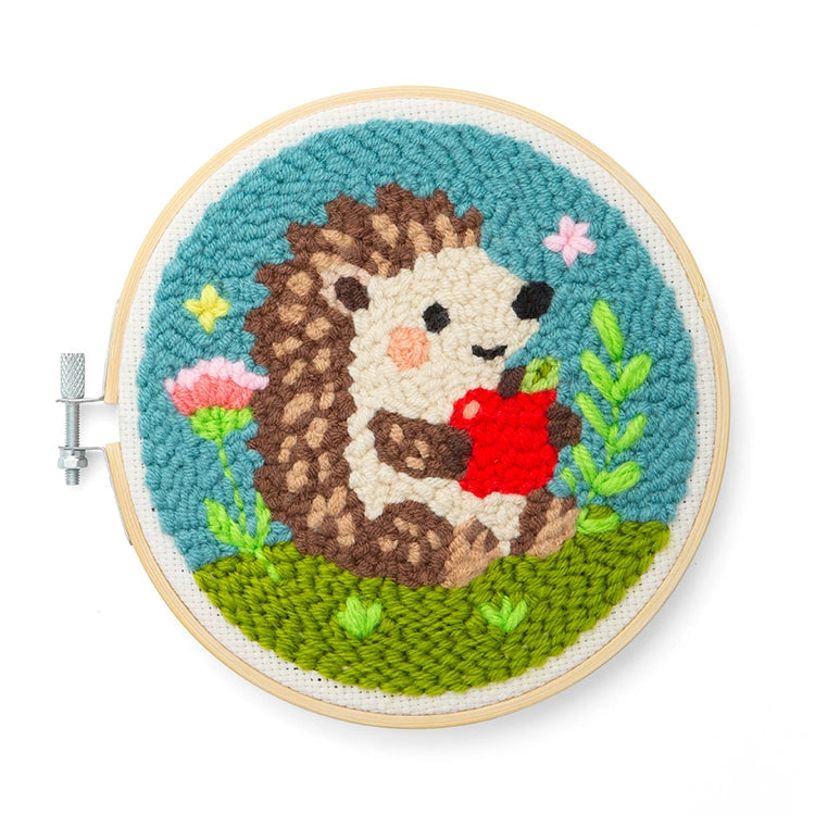 A round wood frame with an iron screw holding a rug hooked sample featuring a hedgehog holding an apple surrounded by flowers, green grass and a blue sky. Shown on a white background.