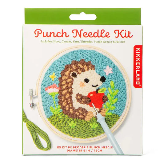 Product pakaging for a 'Punch Needle Kit'. On the front of the packaging is an image of a round wooden frame with a metal screw holding a sample of rug hooking showing a hedgehog holding an apple. All shown on a white background.