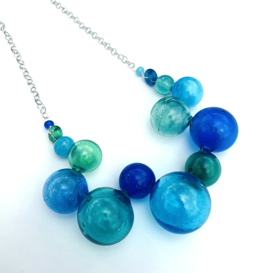 A statement necklace featuring translucent glass beads in various shades of blue, teal, and emerald green, suspended from a silver link chain on a white background.