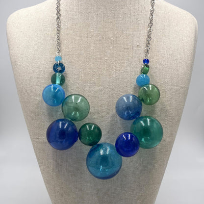 A chunky statement necklace with translucent glass beads in shades of blue, teal, and green, suspended from a silver chain and displayed on a beige fabric mannequin bust.