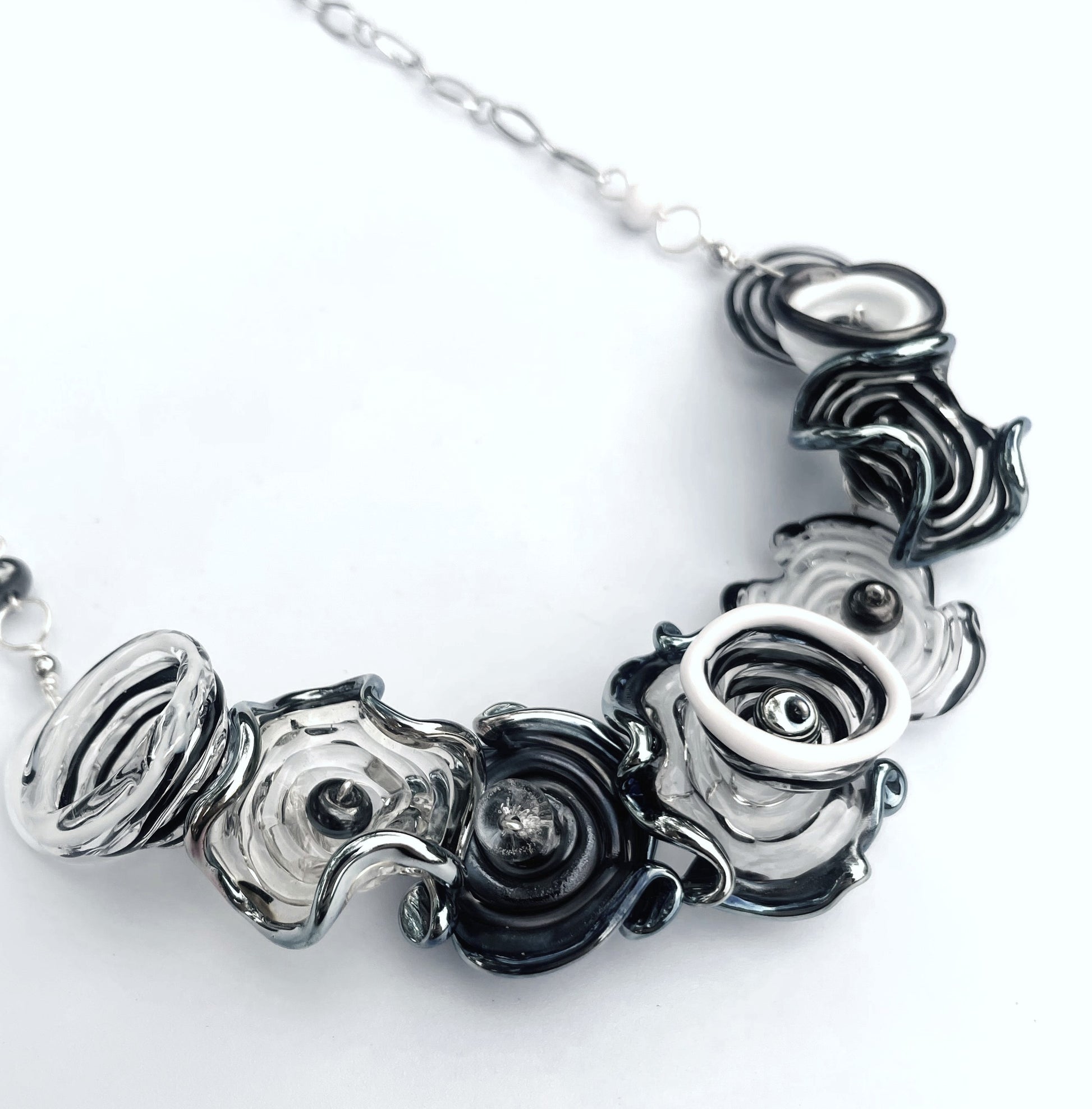 A close-up of a statement necklace featuring textured, sculptural flower-like glass beads in black, white, and smoky gray, suspended from a silver-toned chain against a plain white background.