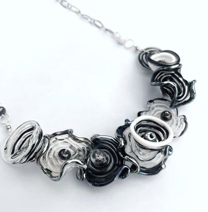 A close-up of a statement necklace featuring textured, sculptural flower-like glass beads in black, white, and smoky gray, suspended from a silver-toned chain against a plain white background.