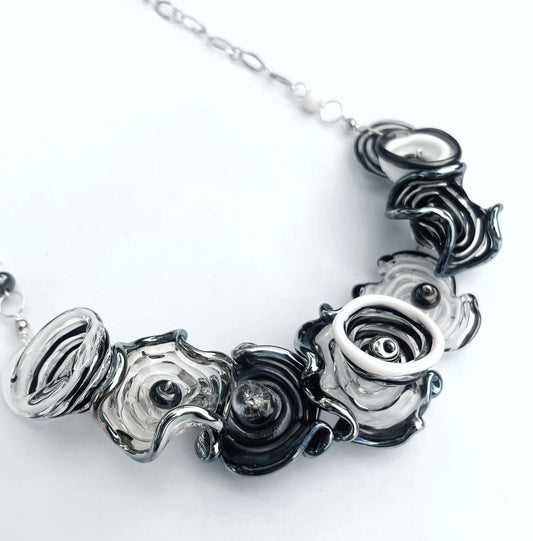 A close-up of a statement necklace featuring textured, sculptural flower-like glass beads in black, white, and smoky gray, suspended from a silver-toned chain against a plain white background.