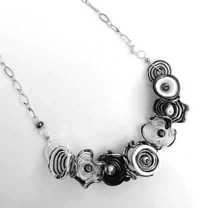A silver-toned statement necklace featuring multiple layered, spiral, and textured flower-like glass beads in black, white, and gray on a plain white background.