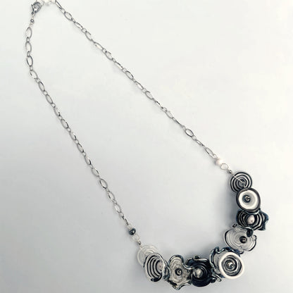 A silver chain necklace featuring a cluster of black and silver spiral flower-shaped glass beads on a plain white background.