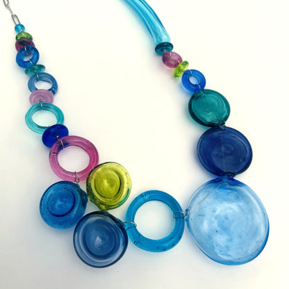 A colorful glass bead necklace featuring large translucent discs in shades of blue, teal, magenta, and lime green, connected by smaller blue and purple beads on a white background.