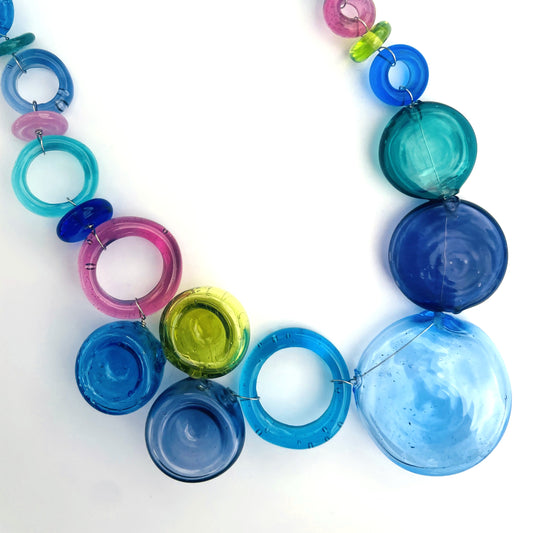 A colorful glass bead necklace featuring translucent disc and ring beads in shades of blue, teal, pink, and lime green, arranged on a plain white background.