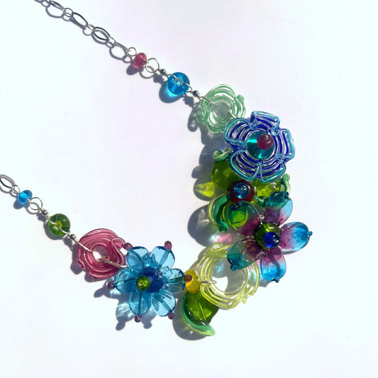 A handmade glass flower necklace featuring multiple translucent petals in shades of blue, green, pink, and purple, attached to a silver-toned metal chain on a white background.