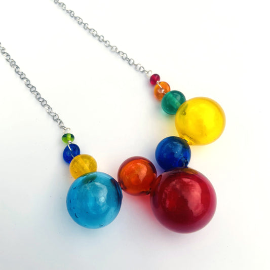A colorful beaded necklace with translucent glass spheres in red, orange, yellow, green, blue, and purple, suspended from a silver chain on a white background.