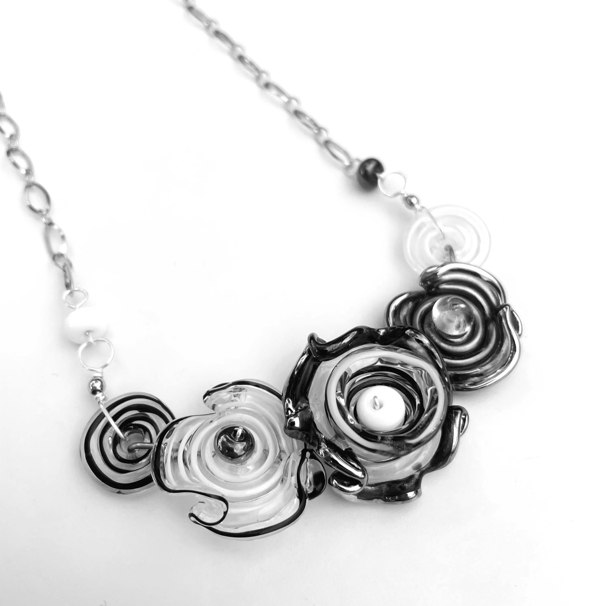 A close-up of a necklace with sculptural, layered spiral flowers in black, white, and clear glass set against a plain white background.