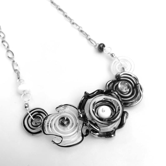 A close-up of a necklace with sculptural, layered spiral flowers in black, white, and clear glass set against a plain white background.