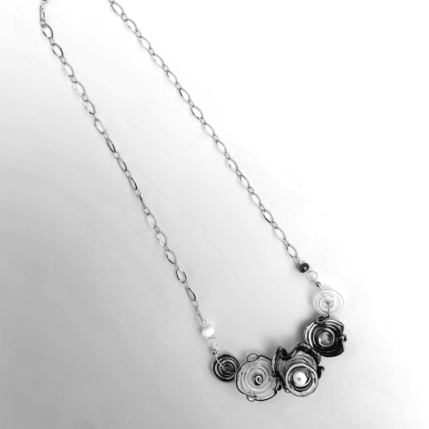 A silver-toned chain necklace featuring a cluster of sculpted, layered glass flowers in black, silver, and white tones, displayed on a light gray background.