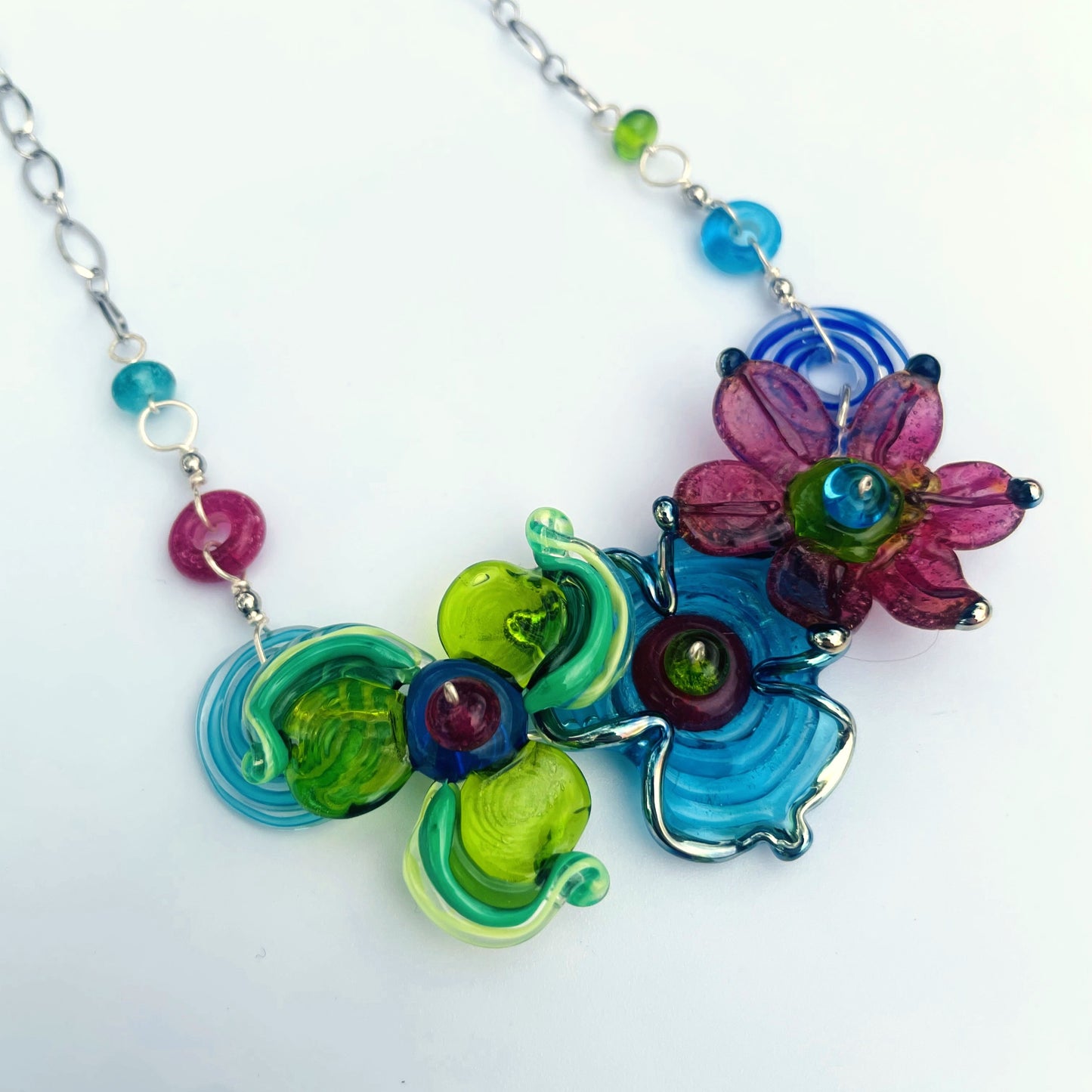 A colorful handmade glass flower necklace on a silver chain, featuring vibrant green, blue, pink, and purple glass petals with small round beads.