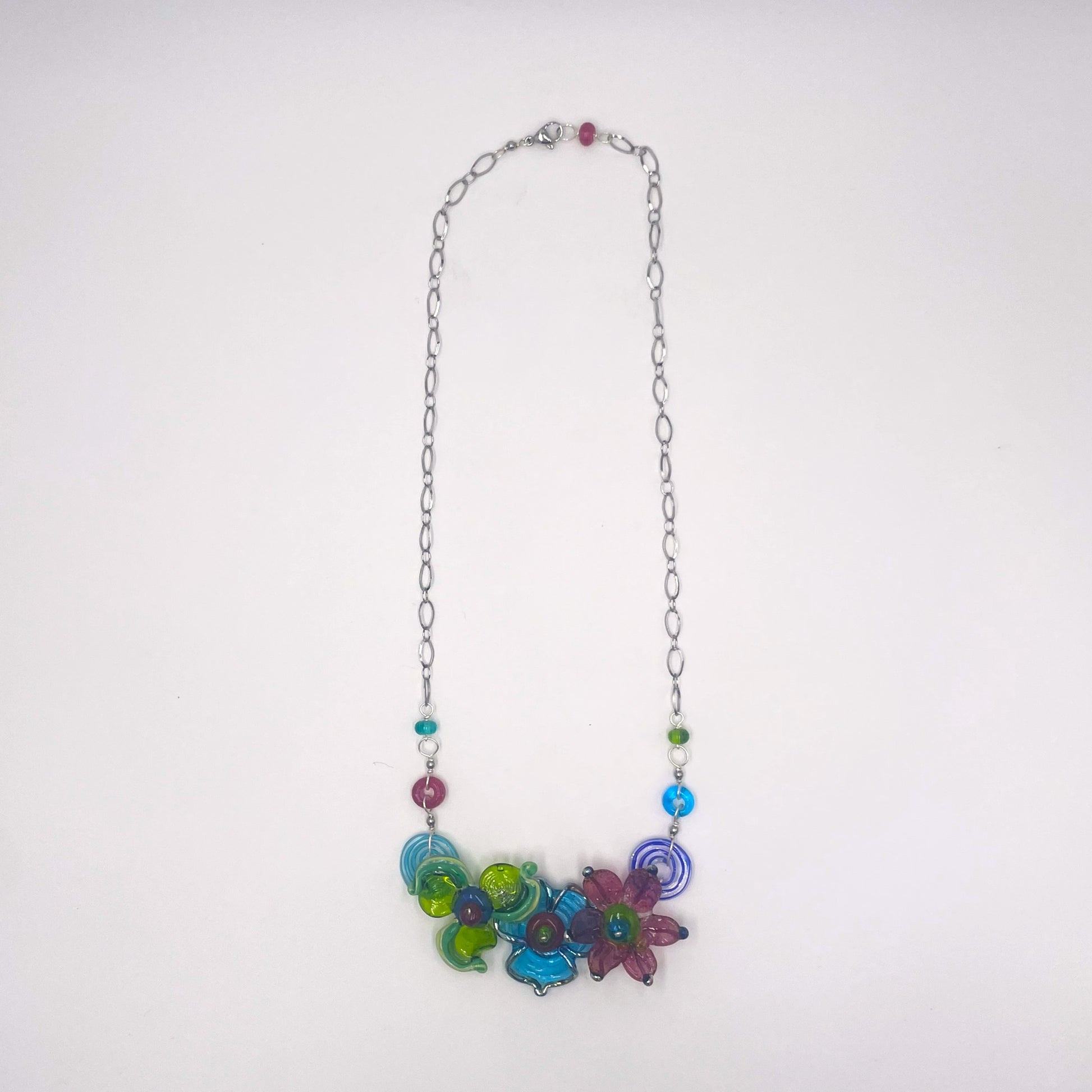 A silver chain necklace featuring a colorful cluster of glass flower charms in shades of blue, green, purple, and pink, accented with small round beads, shown against a plain white background.