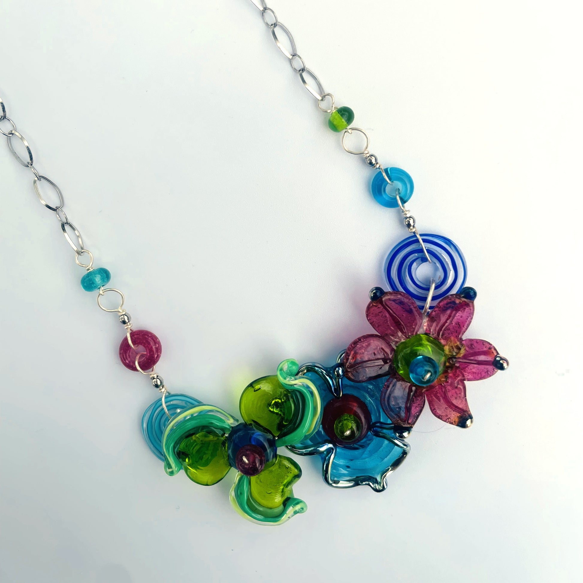 A colorful glass bead and flower statement necklace on a silver chain, featuring translucent green, blue, pink, and purple glass elements on a white background.
