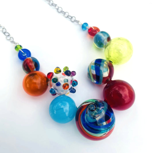 A colorful glass bead necklace featuring a variety of round and marbled beads in red, blue, orange, yellow, and green, with a small decorative bead cluster and a silver chain, shown on a white background.