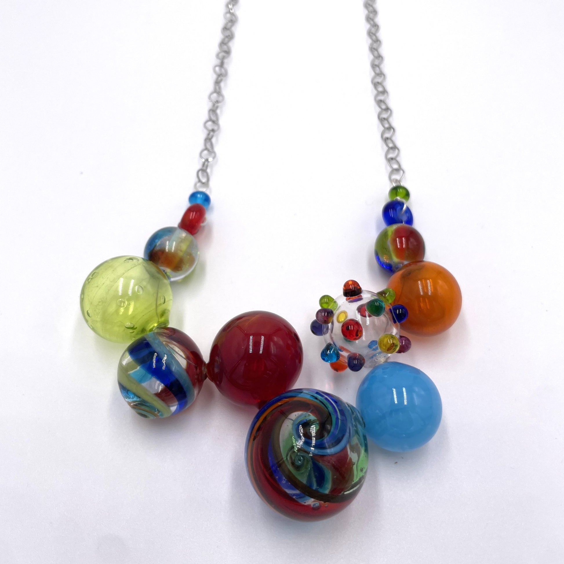 A colorful glass bead necklace with a silver chain, featuring marbled and swirled glass beads in red, blue, green, yellow, orange, and light blue on a white background.