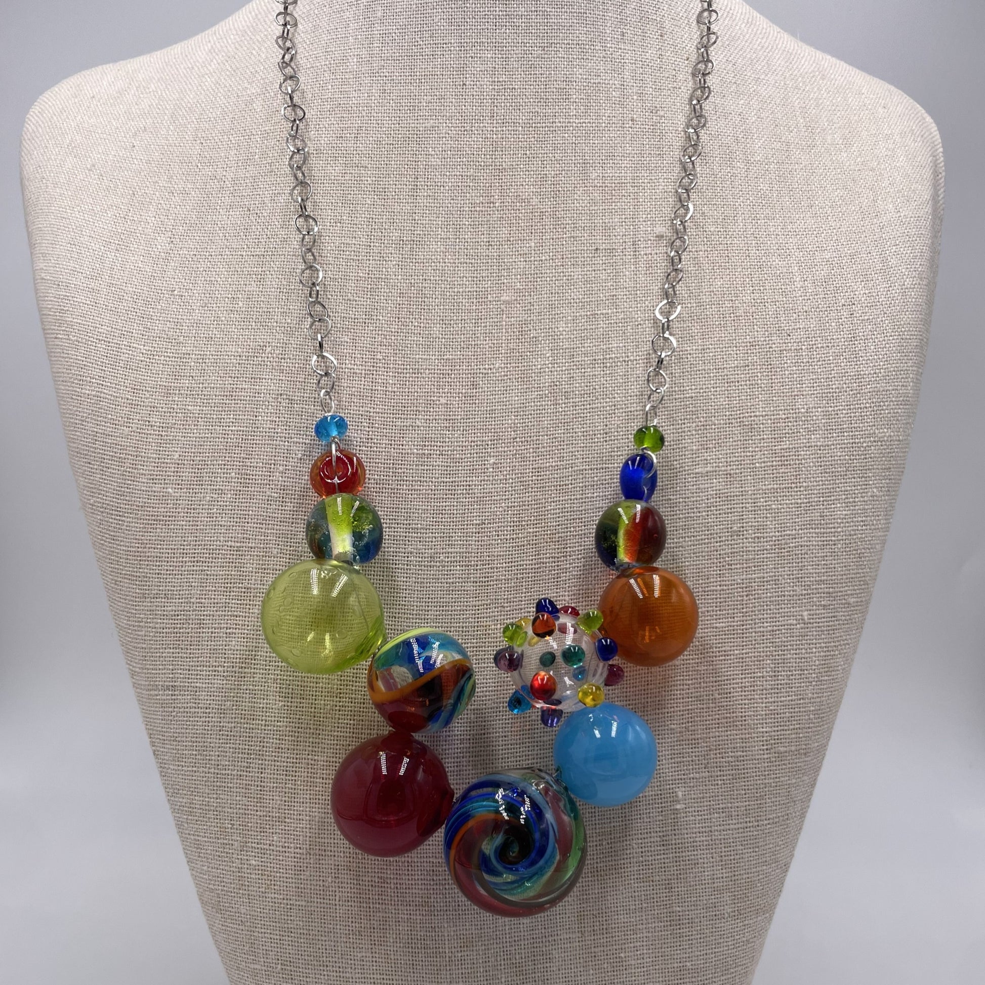 A colorful statement necklace displayed on a beige fabric jewelry bust. The necklace features a silver-tone chain with multiple glass beads in vibrant colors including green, orange, blue, red, and swirl patterns.