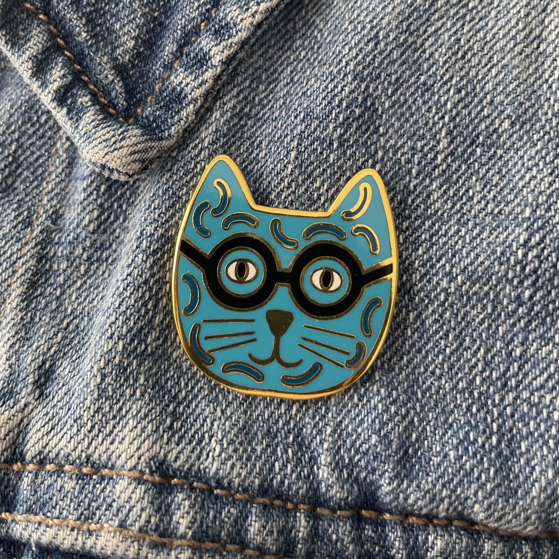 A blue enamel cat pin with black glasses on a blue jean jacket
