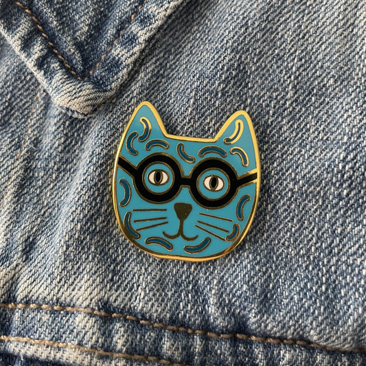 A blue enamel cat pin with black glasses on a blue jean jacket
