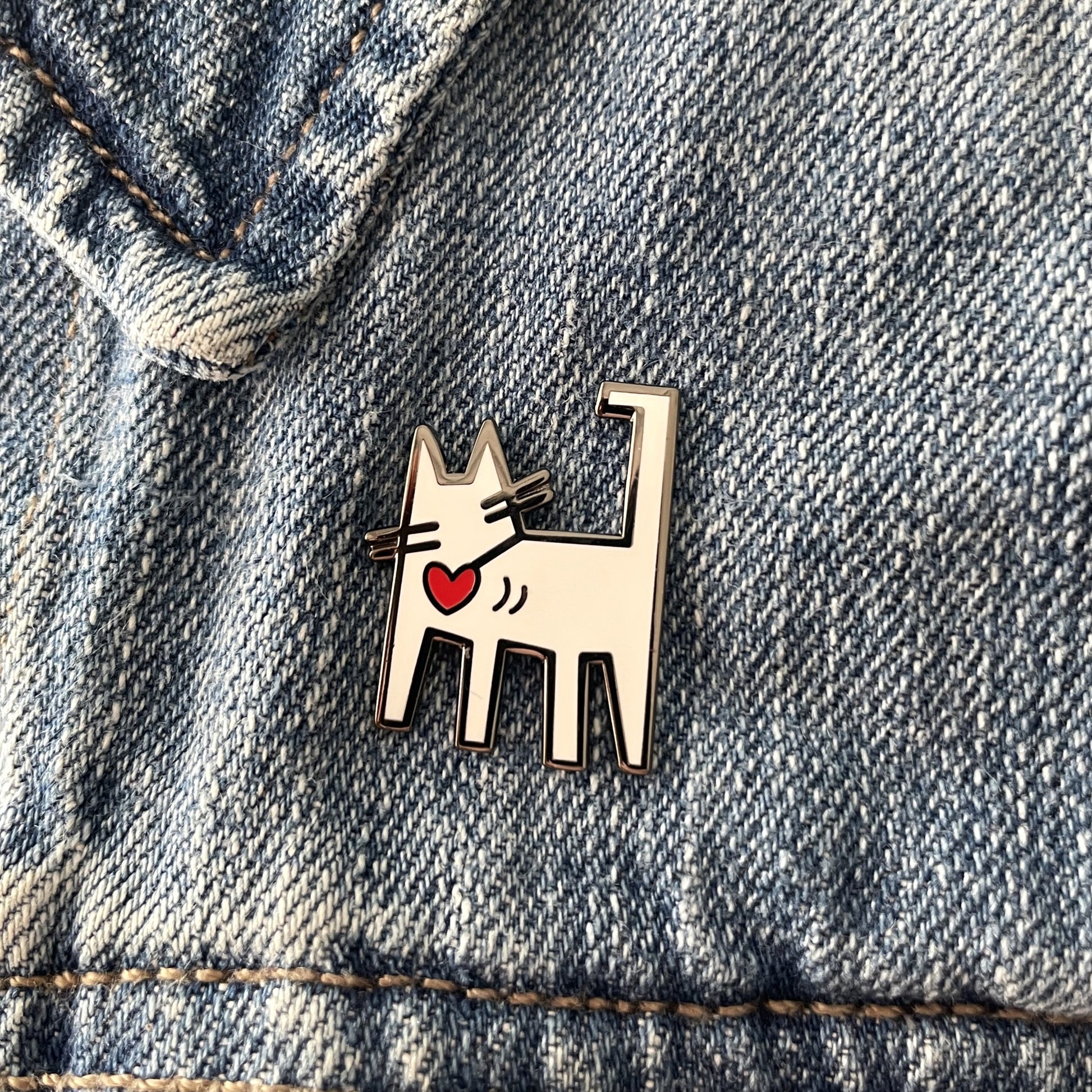 A white enamel cat pin with a red heart on a blue jean jacket