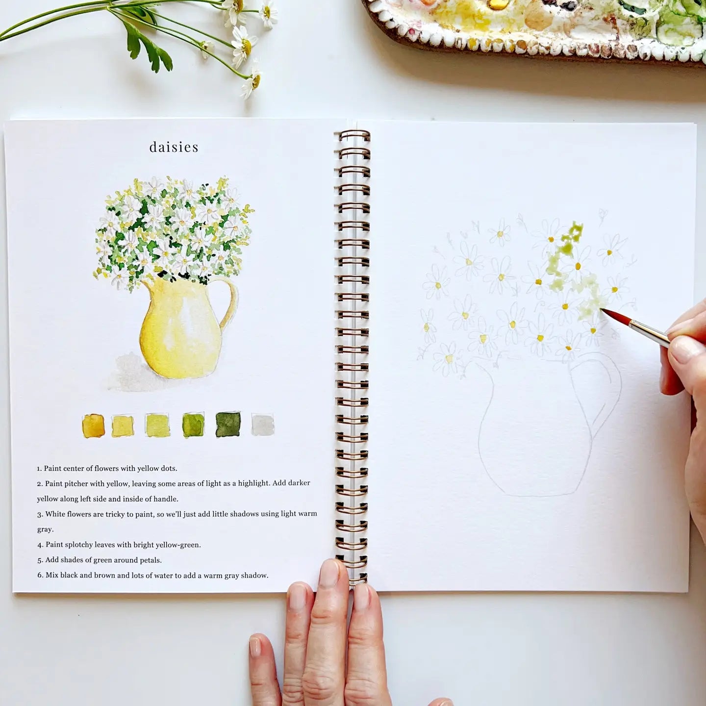 Open notebook with art lesson on daisies, hand holding a paintbrush, and a partially painted daisy.
